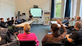 Personen in einem Konferenzraum während einer Präsentation. Einige heben die Hand, ein Banner zur "KI-Ideenwerkstatt für Umweltschutz" steht im Raum.