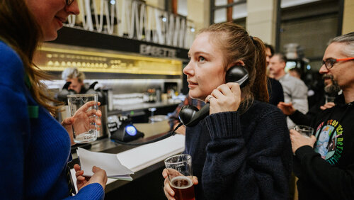 Weiblich gelesene Person hält sich Telefonhörer ans Ohr, im Hintergrund die Theke einer Bar