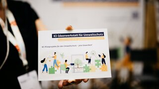 Eine Hand hält eine Postkarte mit Informationen über die KI-Ideenwerkstatt dicht vor die Kamera.